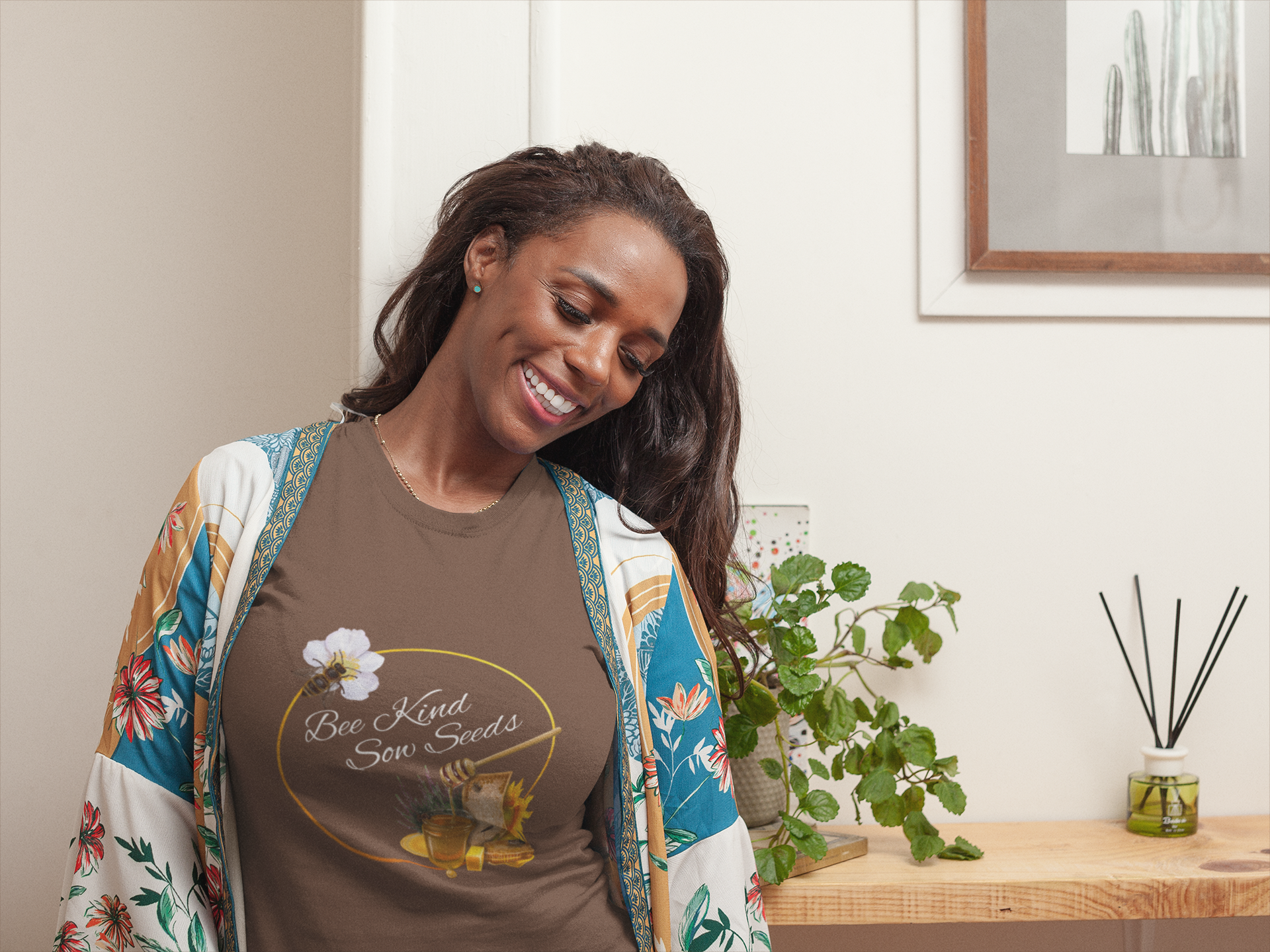 Smiling woman wearing a Bee Kind Sow Seeds in white text on brown shirt with honey, a bee, and a golden circle.